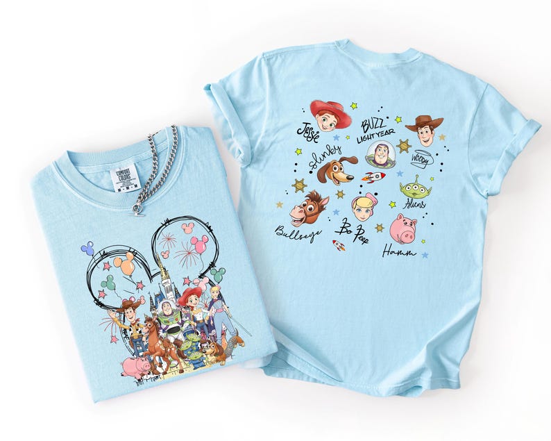 K&ouml;nnte beinhalten: Zwei hellblaue T-Shirts mit Toy Story-Charakteren. Ein Shirt hat ein Mickey Mouse-Ohren-Design mit Feuerwerk und Charakteren. Das andere Shirt zeigt Gesichter von Charakteren und Namen wie Buzz Lightyear und Woody.