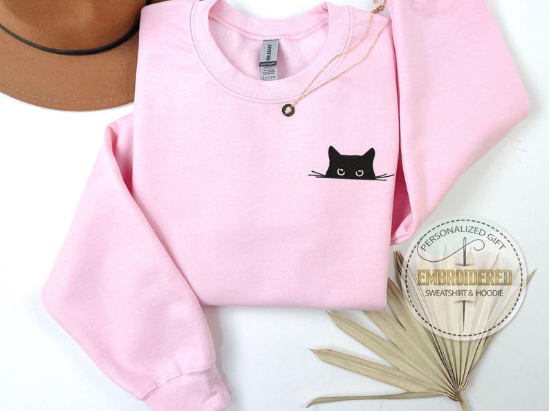 Op de afbeelding: Een roze sweatshirt met een zwarte kat die van onderuit het shirt kijkt. De kat heeft een simpel ontwerp met zwarte ogen en snorharen.