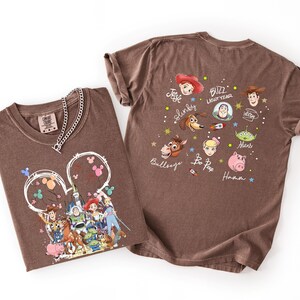 K&ouml;nnte beinhalten: Zwei braune T-Shirts mit Toy Story-Charakteren von Disney. Ein Shirt zeigt eine Grafik von Charakteren vor einem Schloss, das andere hat Gesichter und Namen von Charakteren. Eine silberne Halskette befindet sich auf dem ersten Shirt.