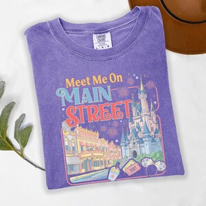 May include: A purple Comfort Colors t-shirt featuring a colorful graphic with the text "Meet Me On Main Street". The design includes a castle, buildings, fireworks, and other theme park elements. The shirt is a size L-G-G.