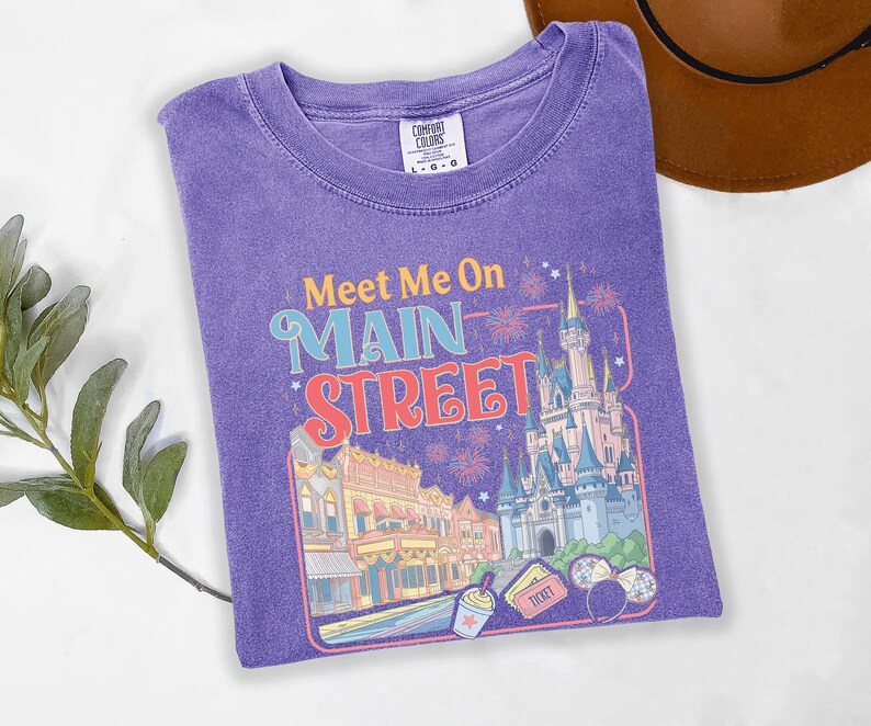 May include: A purple Comfort Colors t-shirt featuring a colorful graphic with the text "Meet Me On Main Street". The design includes a castle, buildings, fireworks, and other theme park elements. The shirt is a size L-G-G.
