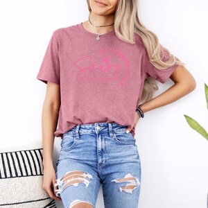 May include: A woman wearing a mauve t-shirt with a pink graphic that says "I like my coffee black and my tea strong". She is wearing blue jeans with distressed details.