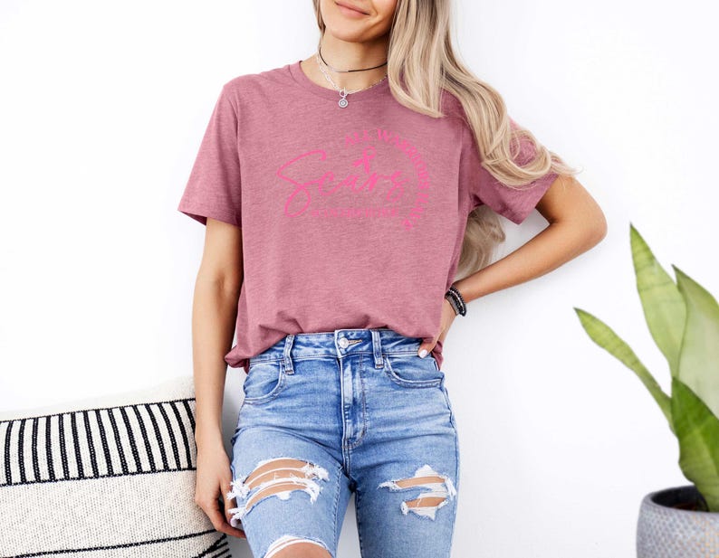 May include: A woman wearing a mauve t-shirt with a pink graphic that says "I like my coffee black and my tea strong". She is wearing blue jeans with distressed details.