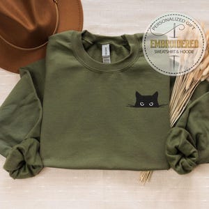 Op de afbeelding: Een groen sweatshirt met lange mouwen met een geborduurd zwart kattengezicht op de voorkant. De kat kijkt naar rechts met open ogen. Het sweatshirt ligt op een lichtbruin oppervlak.