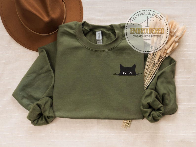 Op de afbeelding: Een groen sweatshirt met lange mouwen met een geborduurd zwart kattengezicht op de voorkant. De kat kijkt naar rechts met open ogen. Het sweatshirt ligt op een lichtbruin oppervlak.