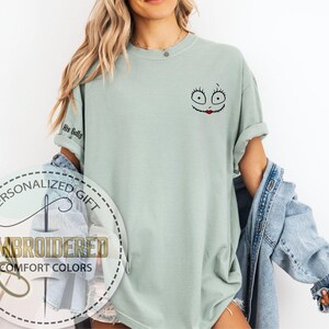 May include: A pale green t-shirt with an embroidered cartoon face on the upper right chest. The design features large eyes and a red stitched mouth. The left sleeve has the text "His Sally". The shirt is part of a personalized gift collection.