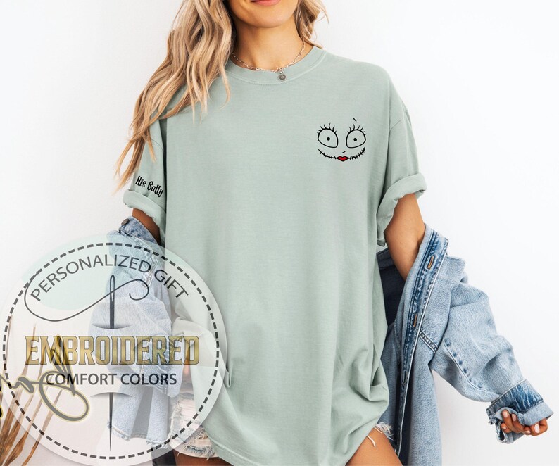 May include: A pale green t-shirt with an embroidered cartoon face on the upper right chest. The design features large eyes and a red stitched mouth. The left sleeve has the text "His Sally". The shirt is part of a personalized gift collection.