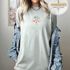 Op de afbeelding: Een lichtgroen T-shirt met een eenvoudige lijntekening van Mickey Mouse in rood, geel en zwart. Het T-shirt wordt gedragen door een persoon met lang blond haar en een spijkerjack.