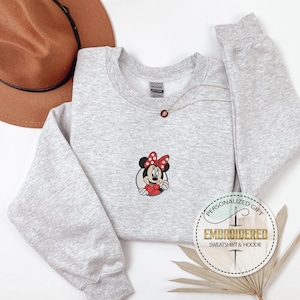 May include: A gray sweatshirt with a red and black embroidered image of Minnie Mouse. The sweatshirt has a crew neck and long sleeves. The text "PERSONALIZED GIFT EMBROIDERED SWEATSHIRT & HOODIE" is on a white circle with a brown stitched border.