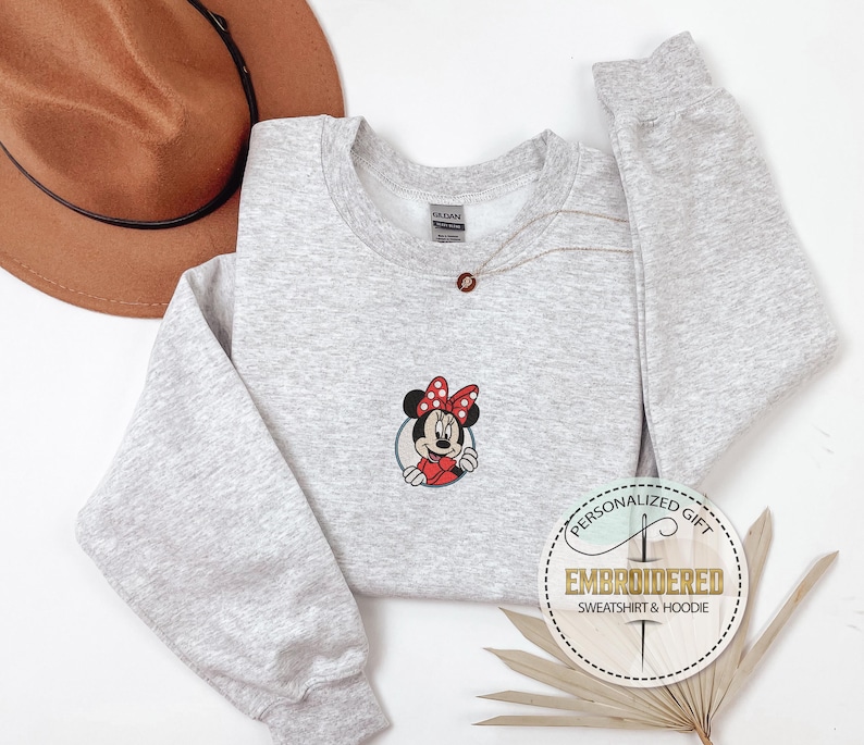 May include: A gray sweatshirt with a red and black embroidered image of Minnie Mouse. The sweatshirt has a crew neck and long sleeves. The text "PERSONALIZED GIFT EMBROIDERED SWEATSHIRT & HOODIE" is on a white circle with a brown stitched border.