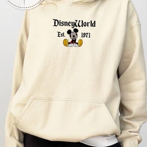 May include: A cream-coloured hoodie with a front pocket and drawstring. The front displays the text "DisneyWorld" above "Est. 1971" and a Mickey Mouse graphic. The hoodie is part of a personalized gift collection.