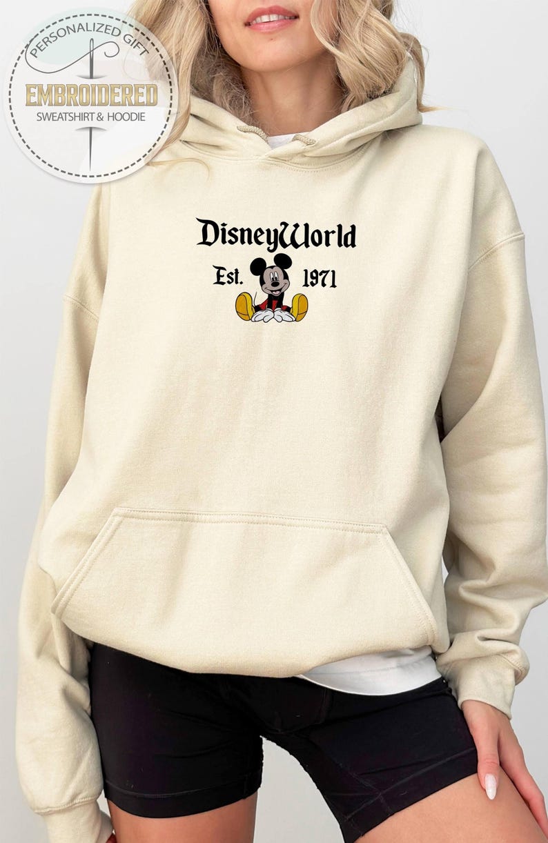 May include: A cream-coloured hoodie with a front pocket and drawstring. The front displays the text "DisneyWorld" above "Est. 1971" and a Mickey Mouse graphic. The hoodie is part of a personalized gift collection.