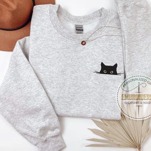 Op de afbeelding: Een grijs sweatshirt met een zwarte geborduurde kat die uit de stof gluurt. Het sweatshirt heeft een ronde hals en lange mouwen. De tekst "Gepersonaliseerd cadeau" en "Geborduurd sweatshirt & hoodie" is zichtbaar in de rechterbenedenhoek van de afbeelding.