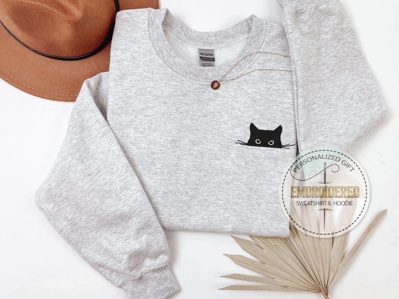 Op de afbeelding: Een grijs sweatshirt met een zwarte geborduurde kat die uit de stof gluurt. Het sweatshirt heeft een ronde hals en lange mouwen. De tekst "Gepersonaliseerd cadeau" en "Geborduurd sweatshirt & hoodie" is zichtbaar in de rechterbenedenhoek van de afbeelding.