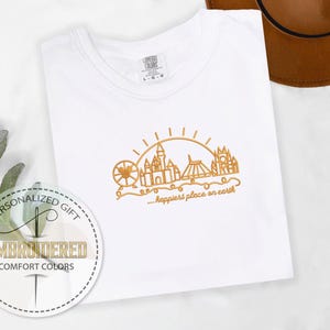 May include: White Comfort Colors t-shirt with gold embroidered design of a Ferris wheel, castle, and other landmarks under a sun. The text "...happiest place on earth" is below the design. A personalized gift tag is in the lower left.