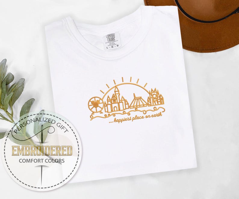 May include: White Comfort Colors t-shirt with gold embroidered design of a Ferris wheel, castle, and other landmarks under a sun. The text "...happiest place on earth" is below the design. A personalized gift tag is in the lower left.