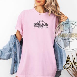 May include: A light pink t-shirt with a black graphic of a cityscape, Ferris wheel, and sun. The text "...happiest place on earth" is below the graphic. The shirt has a relaxed fit and is part of the "Comfort Colors" collection.