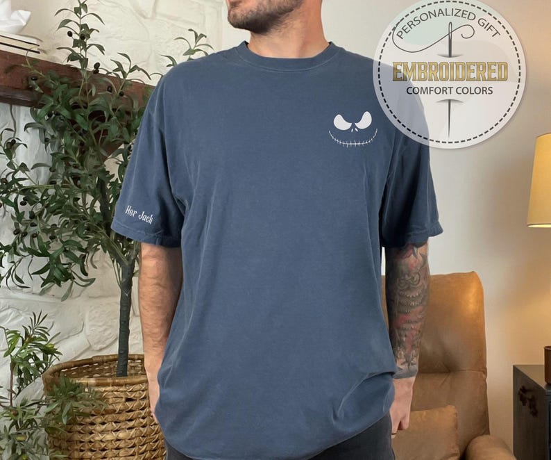 May include: A navy blue t-shirt with a white embroidered Jack Skellington face on the chest and "Her Jack" on the sleeve. The shirt also features a circular logo that says "Personalized Gift" and "Embroidered Comfort Colors".