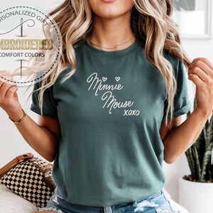 May include: Teal-colored t-shirt with the words "Minnie Mouse xoxo" in white script. The shirt also features a circular logo with the words "Personalized Gift," "Embroidered," and "Comfort Colors."