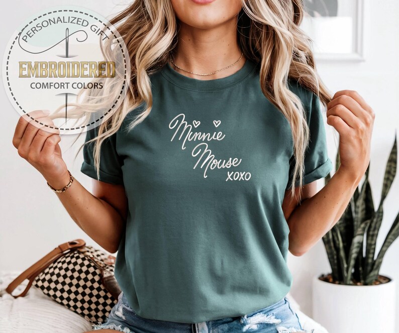 May include: Teal-colored t-shirt with the words "Minnie Mouse xoxo" in white script. The shirt also features a circular logo with the words "Personalized Gift," "Embroidered," and "Comfort Colors."