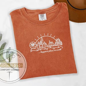 May include: An orange Comfort Colors t-shirt with a white graphic of a Ferris wheel, castle, and other structures, with the text "...happiest place on earth." The shirt is folded and has a tag in the upper right corner.