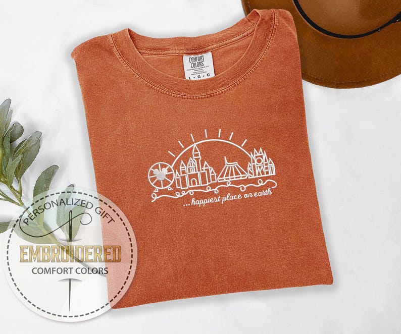 May include: An orange Comfort Colors t-shirt with a white graphic of a Ferris wheel, castle, and other structures, with the text "...happiest place on earth." The shirt is folded and has a tag in the upper right corner.