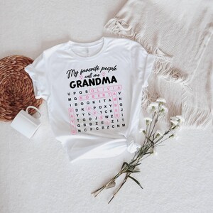 May include: White t-shirt with the text "My favorite people call me Grandma" in black script. A word search grid highlights names like Olivia and Robert. The shirt is knotted at the waist, with a mug and flowers nearby.