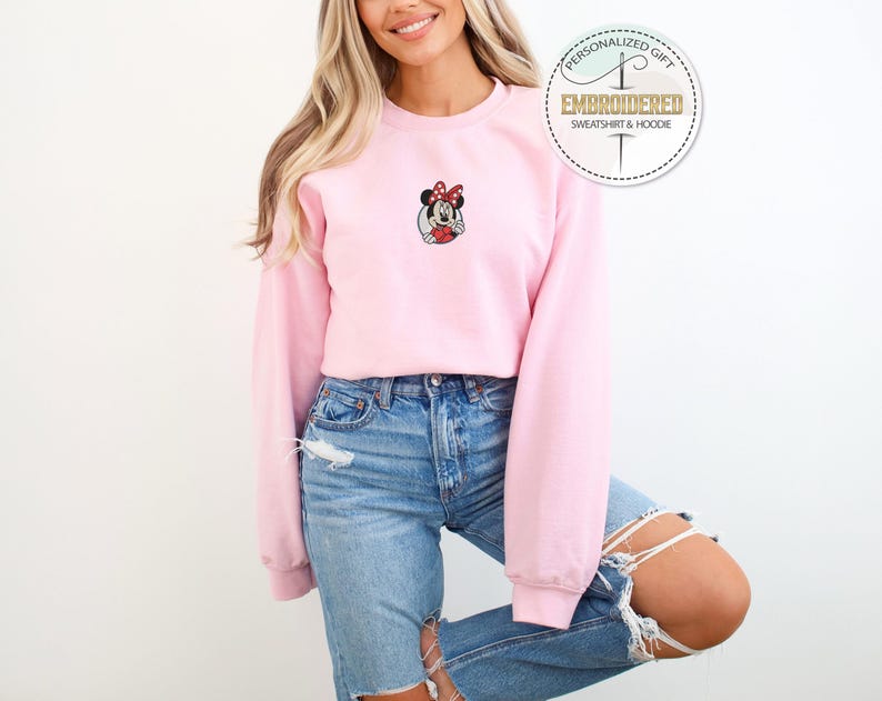 May include: A pink sweatshirt with a cartoon image of Minnie Mouse embroidered on the front. The sweatshirt has long sleeves and a crew neck.