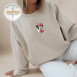 May include: A light beige sweatshirt with a small embroidered image of Mickey Mouse's girlfriend Minnie Mouse. The embroidered image shows Minnie Mouse with her signature red bow and white polka dots.