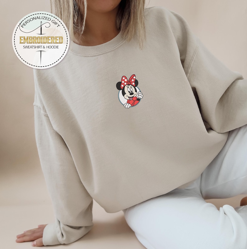 May include: A light beige sweatshirt with a small embroidered image of Mickey Mouse's girlfriend Minnie Mouse. The embroidered image shows Minnie Mouse with her signature red bow and white polka dots.
