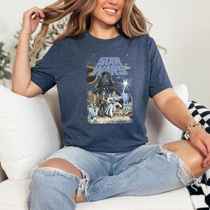 May include: Navy blue t-shirt featuring a Star Wars movie poster design. The graphic includes Darth Vader, other characters, and the Star Wars logo in blue and yellow. The shirt is worn by a person.
