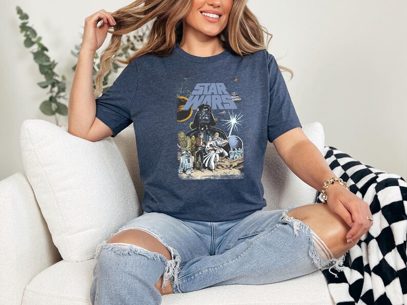 May include: Navy blue t-shirt featuring a Star Wars movie poster design. The graphic includes Darth Vader, other characters, and the Star Wars logo in blue and yellow. The shirt is worn by a person.