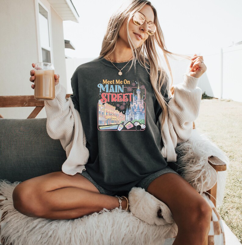 May include: Dark gray sweatshirt with the text "Meet Me On Main Street" in colorful lettering. The graphic features a castle, fireworks, and other park elements. The sweatshirt is worn with shorts and a cream-colored cardigan.