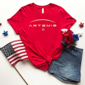 May include: A red t-shirt with the text "ARTEMIS II" in white, paired with a denim skirt, American flag, and star-shaped decorations. The image has a patriotic theme.
