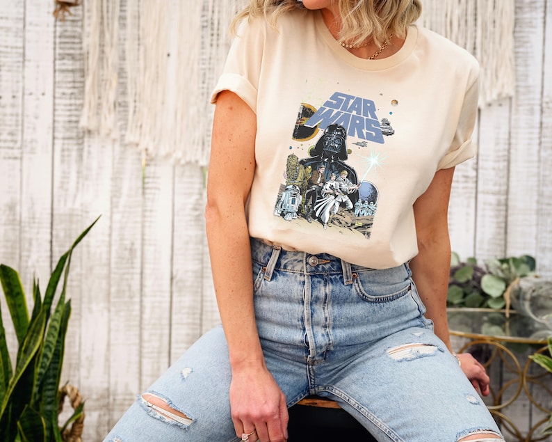 May include: Cream-colored Star Wars graphic tee featuring Darth Vader and other characters. The shirt has a classic crew neck and short sleeves. The graphic is in shades of blue, black, and beige. The model is wearing light blue ripped jeans.