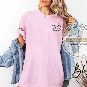 May include: A pale pink t-shirt featuring an embroidered Sally face design on the chest. The sleeve displays the text "His Sally". The shirt is part of a personalized gift collection and is made of comfort colors.