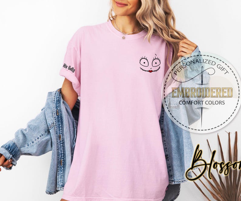May include: A pale pink t-shirt featuring an embroidered Sally face design on the chest. The sleeve displays the text "His Sally". The shirt is part of a personalized gift collection and is made of comfort colors.