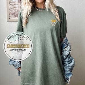 Pu&ograve; includere: Una maglietta verde con un ricamo di fiori gialli sul petto sinistro. La maglietta ha una vestibilit&agrave; rilassata e un collo rotondo. L'immagine include anche un logo per un negozio di regali personalizzati.
