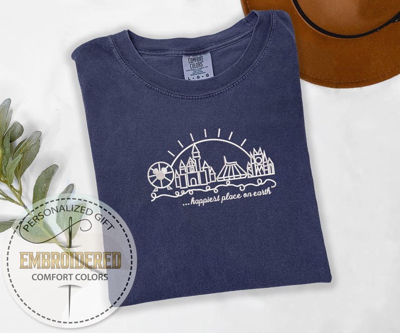 May include: Navy blue Comfort Colors t-shirt with a white line drawing of a theme park skyline, including a Ferris wheel, castle, and other structures. The text "...happiest place on earth" is below the drawing. The shirt is folded on a white surface.