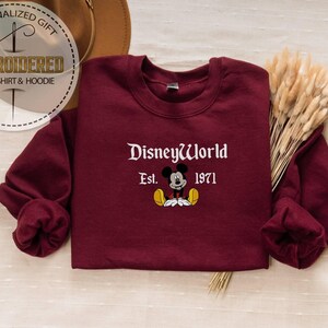 May include: Burgundy sweatshirt featuring "DisneyWorld Est. 1971" and a Mickey Mouse graphic. The long-sleeved sweatshirt has a crew neck. A personalized gift tag is in the upper left corner.