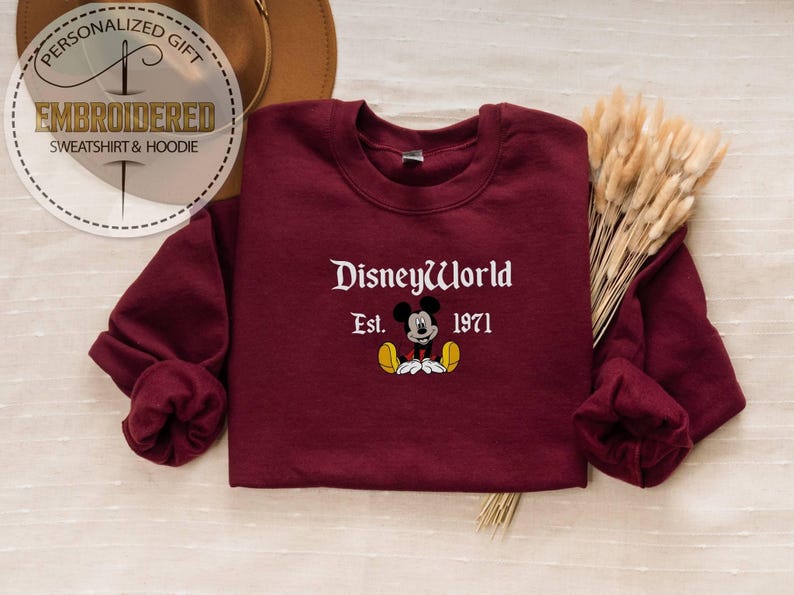 May include: Burgundy sweatshirt featuring "DisneyWorld Est. 1971" and a Mickey Mouse graphic. The long-sleeved sweatshirt has a crew neck. A personalized gift tag is in the upper left corner.