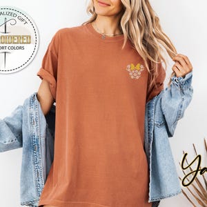 Pu&ograve; includere: Una maglietta color ruggine con un piccolo disegno ricamato di un fiore giallo con accenti bianchi. La maglietta &egrave; indossata da una persona con i capelli lunghi e biondi e una giacca di jeans.