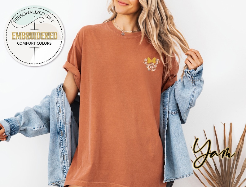 Pu&ograve; includere: Una maglietta color ruggine con un piccolo disegno ricamato di un fiore giallo con accenti bianchi. La maglietta &egrave; indossata da una persona con i capelli lunghi e biondi e una giacca di jeans.