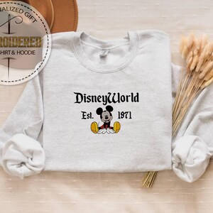 May include: Pale grey sweatshirt featuring the text "DisneyWorld Est. 1971" in black script, with a Mickey Mouse cartoon character beneath. The sweatshirt has long sleeves and a crew neck.