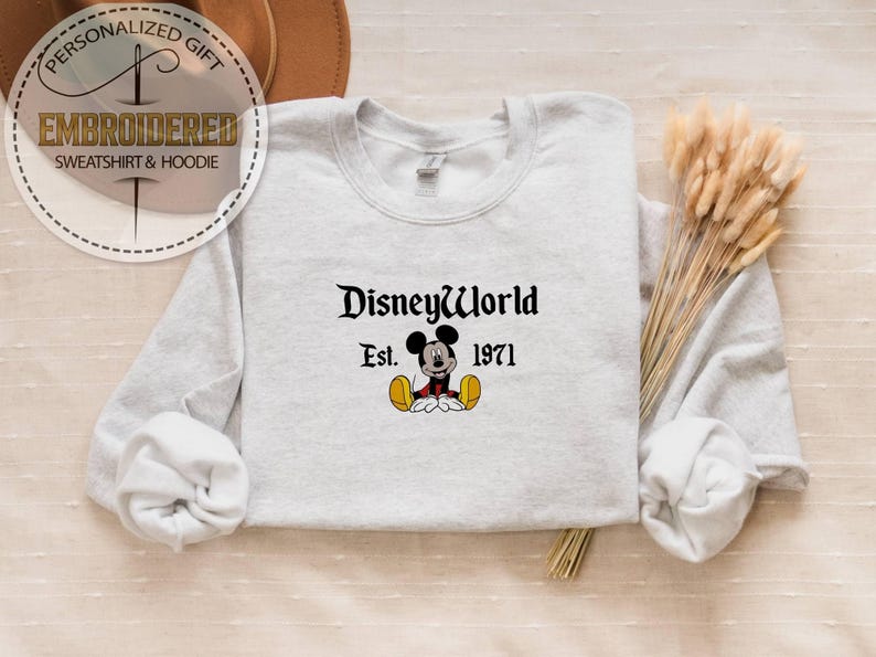 May include: Pale grey sweatshirt featuring the text "DisneyWorld Est. 1971" in black script, with a Mickey Mouse cartoon character beneath. The sweatshirt has long sleeves and a crew neck.
