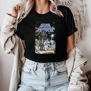 May include: Black t-shirt featuring a Star Wars graphic. The design includes Darth Vader, characters, and the Star Wars logo in blue. The background shows a space scene with planets and stars. A classic movie-themed tee.