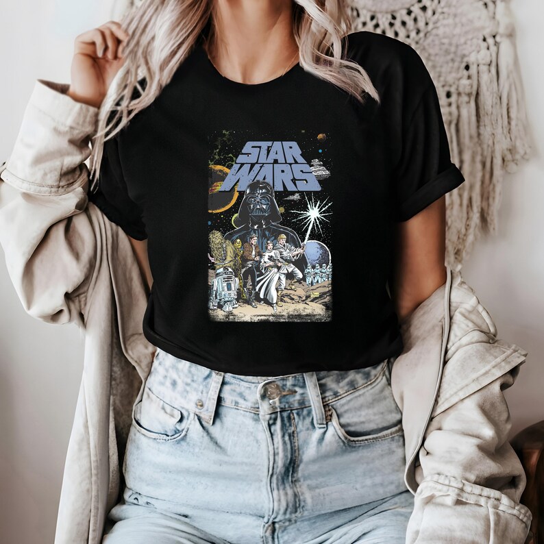 May include: Black t-shirt featuring a Star Wars graphic. The design includes Darth Vader, characters, and the Star Wars logo in blue. The background shows a space scene with planets and stars. A classic movie-themed tee.