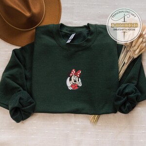 May include: A dark green sweatshirt with a red and white embroidered image of Minnie Mouse. The sweatshirt has a crew neck and long sleeves.