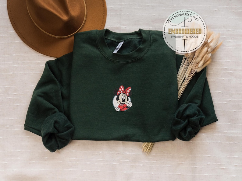 May include: A dark green sweatshirt with a red and white embroidered image of Minnie Mouse. The sweatshirt has a crew neck and long sleeves.