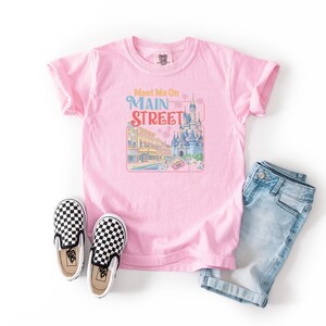 May include: A light pink t-shirt with the words "Meet Me On Main Street" and a colorful graphic of a castle and buildings. The shirt is paired with blue denim shorts and black and white checkered slip-on shoes.